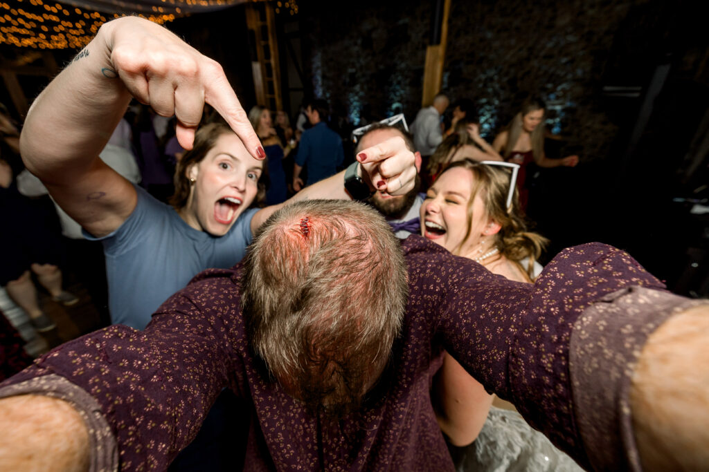 Bride, groom, and second photographer laughing and pointing at the photographer's head staples on the dance floor